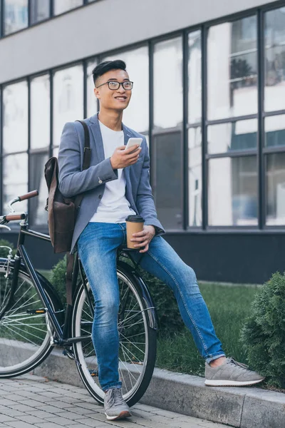 Hombre asiático feliz con taza de café desechable usando teléfono inteligente y sentado en bicicleta - foto de stock