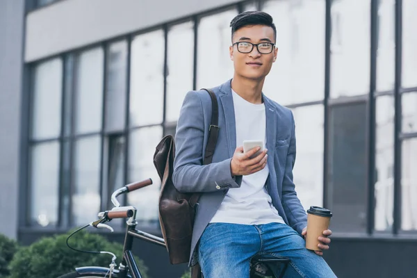 Bel homme asiatique avec café pour aller à l'aide d'un smartphone et assis sur le vélo — Photo de stock