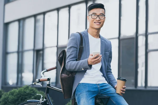 Smiling asian man with coffee to go using smartphone and sitting on bicycle — Stock Photo