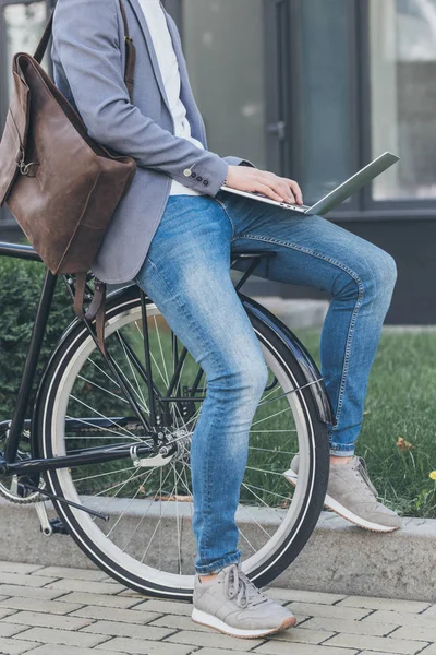 Cropped view of freelancer with leather backpack working on laptop while sitting on bike — Stock Photo
