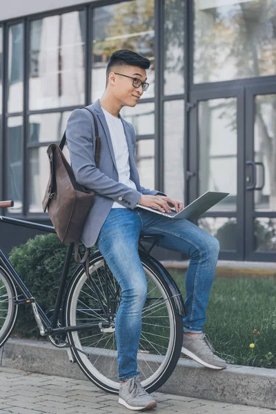 Pensive asian teleworker with leather backpack using laptop while sitting on bicycle — Stock Photo
