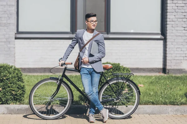 Smiling asian man in trendy jacket holding laptop and standing at bicycle — Stock Photo