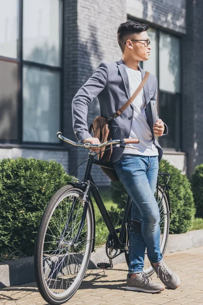 Stylish asian man with laptop and leather bag leaning on bicycle — Stock Photo