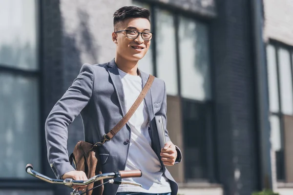 Sourire homme asiatique avec ordinateur portable et sac en cuir debout avec vélo — Photo de stock