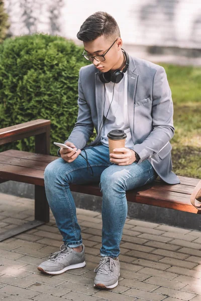 Hombre asiático con estilo con café para llevar, teléfono inteligente y auriculares sentado en el banco - foto de stock