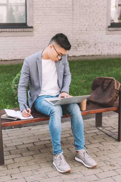 Asiatischer Freiberufler schreibt im Planer, während er auf dem Smartphone spricht und auf der Bank am Laptop arbeitet — Stockfoto