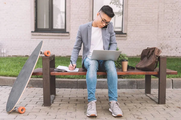 Selbstbewusster asiatischer Telearbeiter schreibt im Planer, während er auf dem Smartphone spricht und Laptop auf Bank mit Longboard benutzt — Stockfoto