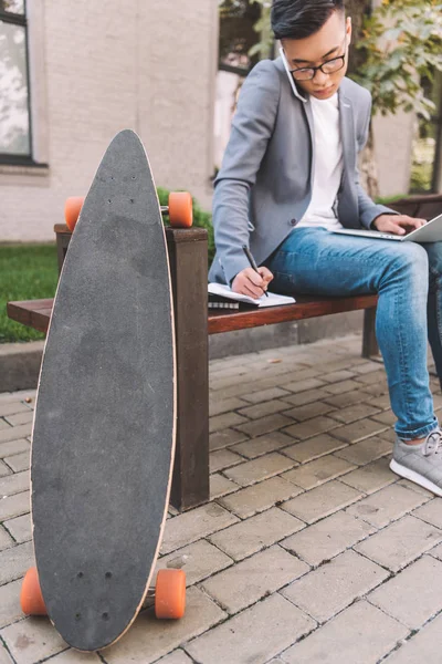 Asian teleworker writing in planner while using smartphone and laptop on bench with longboard — Stock Photo