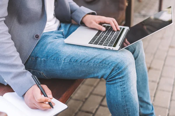 Visão cortada de freelancer trabalhando no laptop e escrevendo em planejador enquanto sentado no banco — Fotografia de Stock