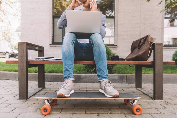Обрезанный вид фрилансера, работающего с ноутбуком и смартфоном, сидя на скамейке с длинной доской — стоковое фото
