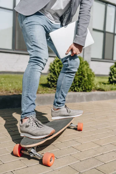 Обрезанный вид стильного человека с ноутбуком скейтбординг на длинной доске — стоковое фото