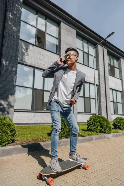 Asian man with laptop having conversation while riding longboard on street — Stock Photo