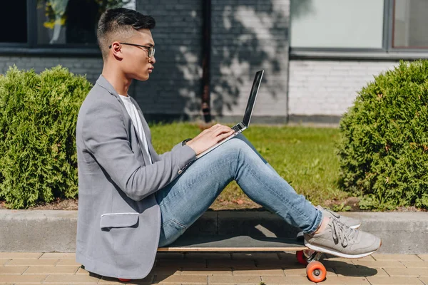 Side view of asian freelancer using laptop while sitting on longboard on street — Stock Photo