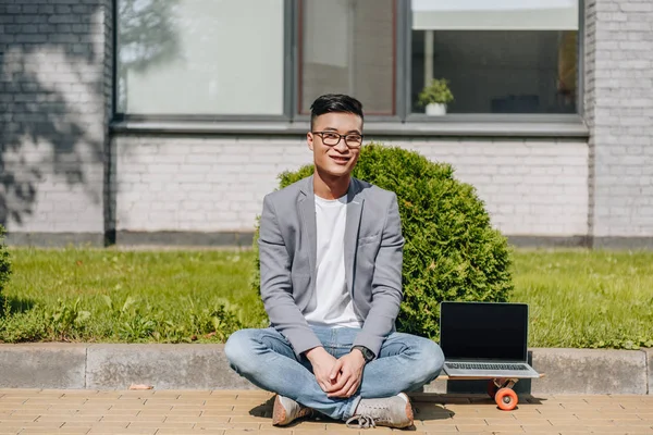 Sourire asiatique homme assis sur longboard avec ordinateur portable avec écran vide sur rue — Photo de stock