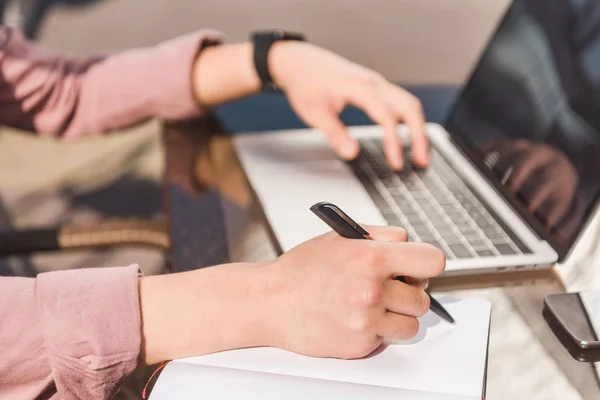 Vue partielle de l'homme travaillant à distance sur un ordinateur portable et prenant des notes sur la table — Photo de stock