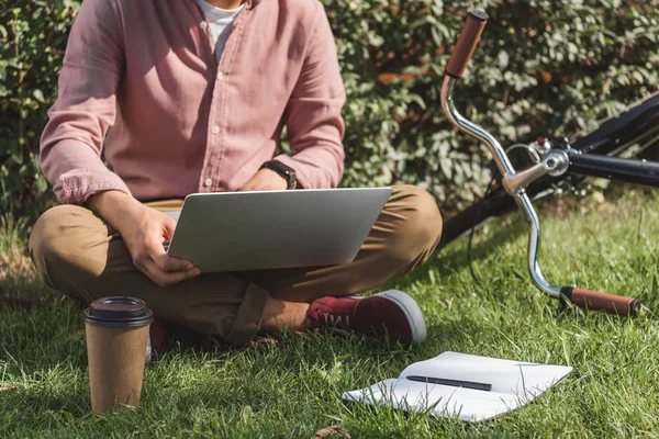 Tiro recortado de freelancer trabalhando no laptop na grama verde com café para ir e notebook no parque — Fotografia de Stock