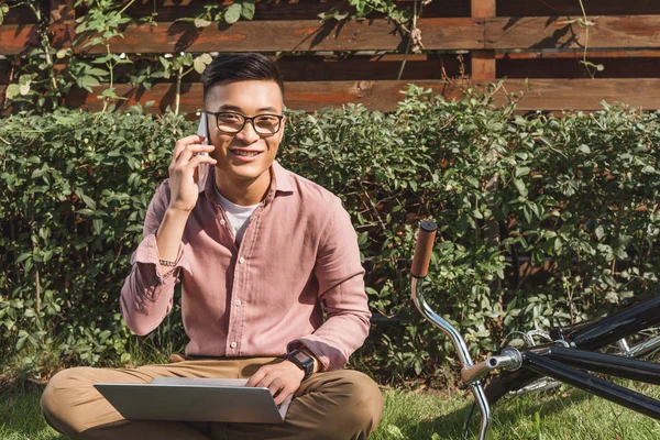 Sourire asiatique homme parler sur smartphone tout en travaillant à distance sur ordinateur portable dans le parc — Photo de stock