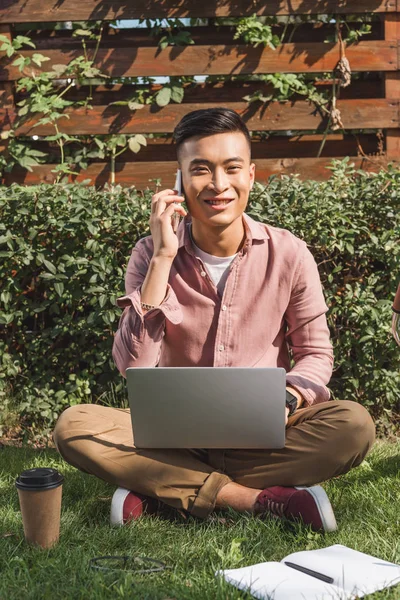 Усміхнений азіатський чоловік говорить на смартфоні, працюючи на ноутбуці в парку — стокове фото
