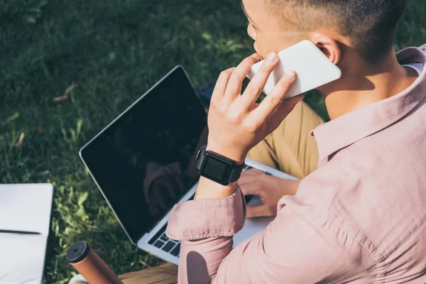 Asian man talking on smartphone while remote working on laptop in park — Stock Photo