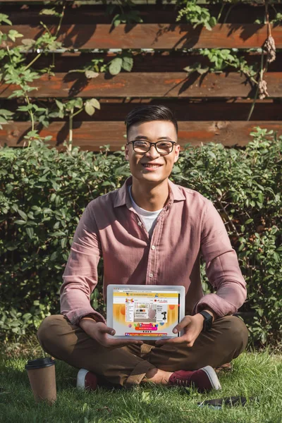 Smiling asian man sitting on green grass and showing tablet with aliexpress website on screen in hands in park — Stock Photo