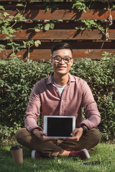 Усміхнений азіатський чоловік сидить на зеленій траві і показує планшет з порожнім екраном в руках в парку — стокове фото