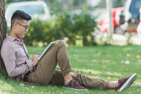 Side view of asian man in eyeglasses using digital tablet on green grass in park — Stock Photo