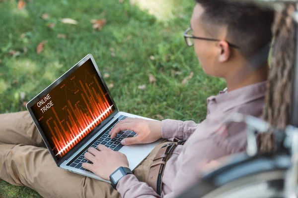 Side view of young asian freelancer working on laptop with online trade lettering in park — Stock Photo