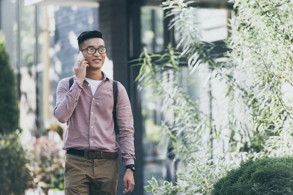 Азиатский мужчина с рюкзаком разговаривает на смартфоне во время прогулки по улице — стоковое фото