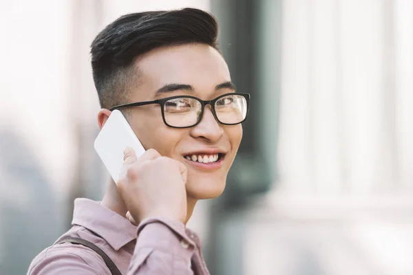 Seitenansicht eines lächelnden asiatischen Mannes mit Brille, der sich auf der Straße mit dem Smartphone unterhält — Stockfoto