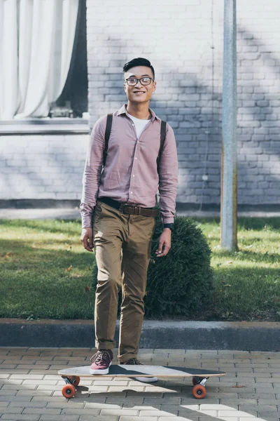 Sonriente asiático hombre con mochila y longboard mirando cámara en calle - foto de stock