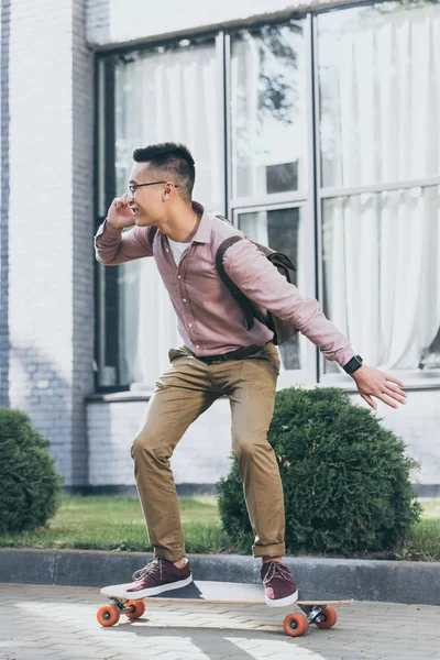 Asian man talking on smartphone while standing on longboard on street — Stock Photo