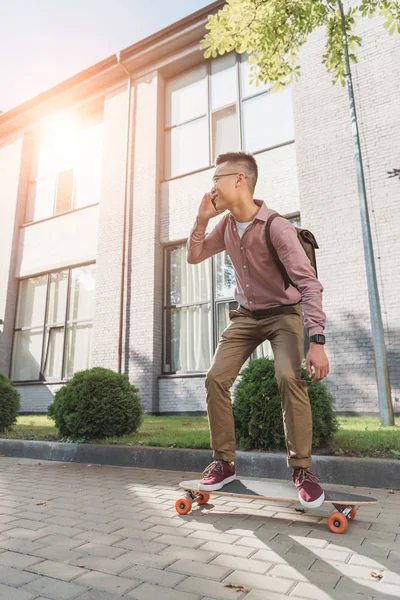 Asian man talking on smartphone while standing on longboard on street — Stock Photo