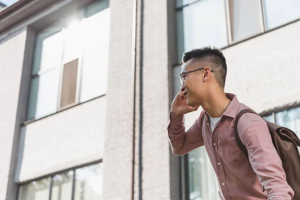 Side view of cheerful asian man in eyeglasses talking on smartphone on street — Stock Photo