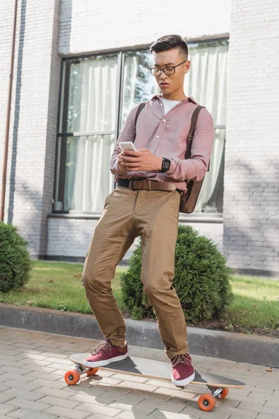 Concentré asiatique l'homme en utilisant smartphone tandis que debout sur longboard sur rue — Photo de stock