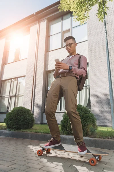Focused asian man using smartphone while standing on longboard on street — Stock Photo