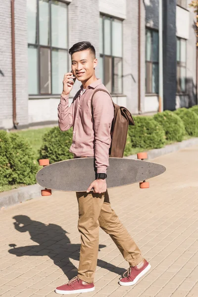 Side view of smiling asian man with longboard having conversation on smartphone on street — Stock Photo