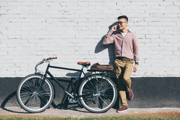 Азиатский мужчина в стильной одежде разговаривает по смартфону, стоя рядом с велосипедом на улице — стоковое фото