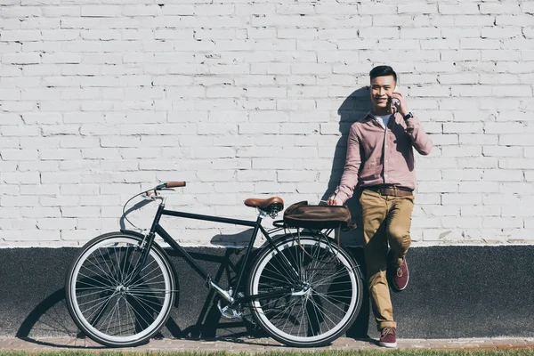 Ásia homem no elegante roupas falando no smartphone no bicicleta no rua — Fotografia de Stock