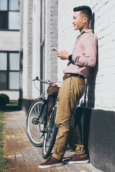 Homem asiático em roupas elegantes com smartphone em pé perto de bicicleta na rua — Fotografia de Stock