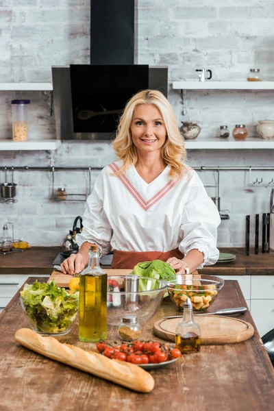 Attraktive lächelnde erwachsene Frau, die zu Hause Salat für das Abendessen zubereitet und in die Kamera schaut — Stockfoto