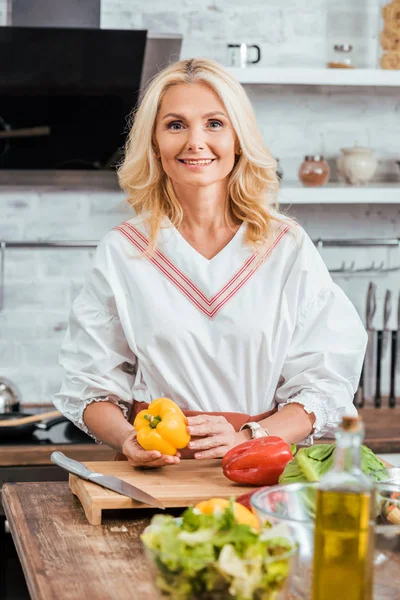 Donna attraente sorridente che prepara l'insalata per cena, tenendo il peperone e guardando la fotocamera a casa — Foto stock