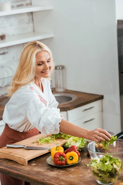 Atractiva mujer preparando ensalada para la cena y poniendo ingredientes en el tazón en casa - foto de stock