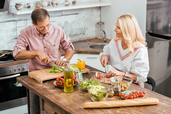 Sorrindo marido e mulher adultos preparando salada para jantar juntos e cortar legumes na cozinha — Fotografia de Stock