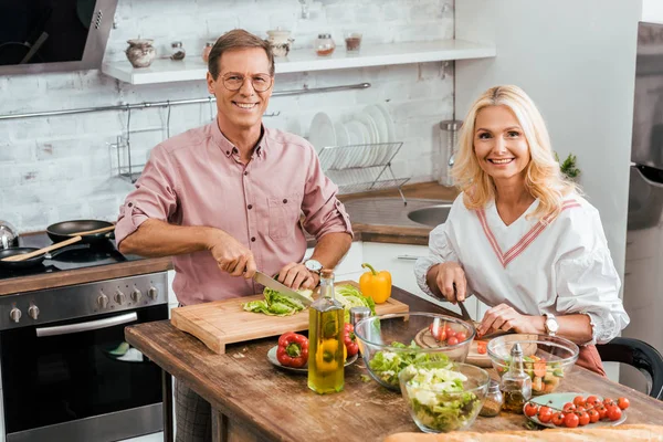 Vue grand angle du couple souriant préparant la salade pour le dîner ensemble dans la cuisine et regardant la caméra — Photo de stock