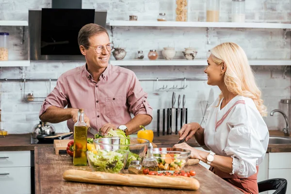 Sorrindo casal preparando salada para jantar juntos na cozinha, olhando um para o outro — Fotografia de Stock