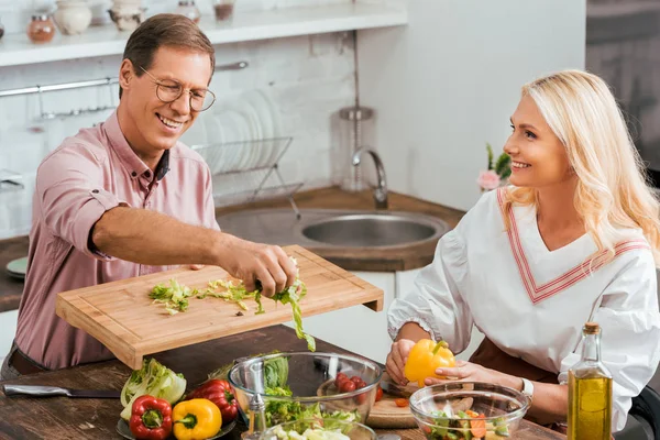 Улыбающаяся пара готовит салат к ужину вместе на кухне — стоковое фото