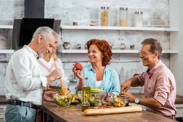 Sorrindo amigos maduros preparando salada para o jantar e conversando em casa — Fotografia de Stock