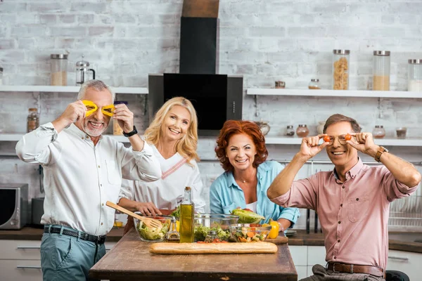 Mature men having fun with vegetables during preparing salad for dinner at home and looking at camera — Stock Photo