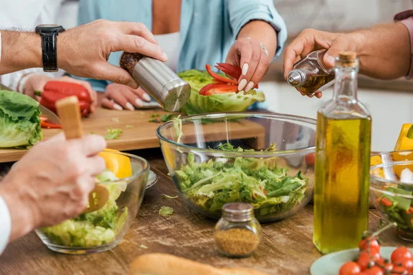 Imagem cortada de amigos maduros preparando salada para o jantar em casa e adicionando especiarias na tigela — Fotografia de Stock