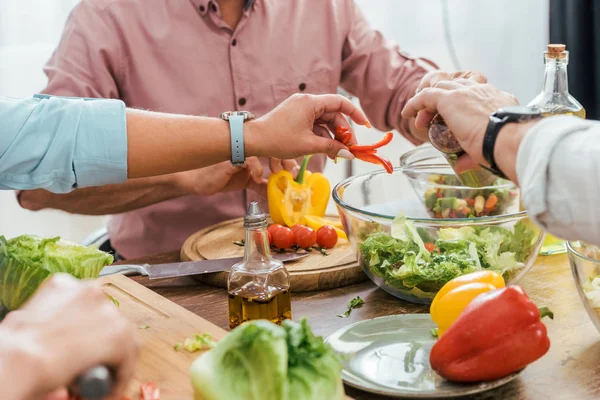 Imagen recortada de amigos maduros preparando ensalada para la cena en casa y poniendo ingredientes en un tazón - foto de stock